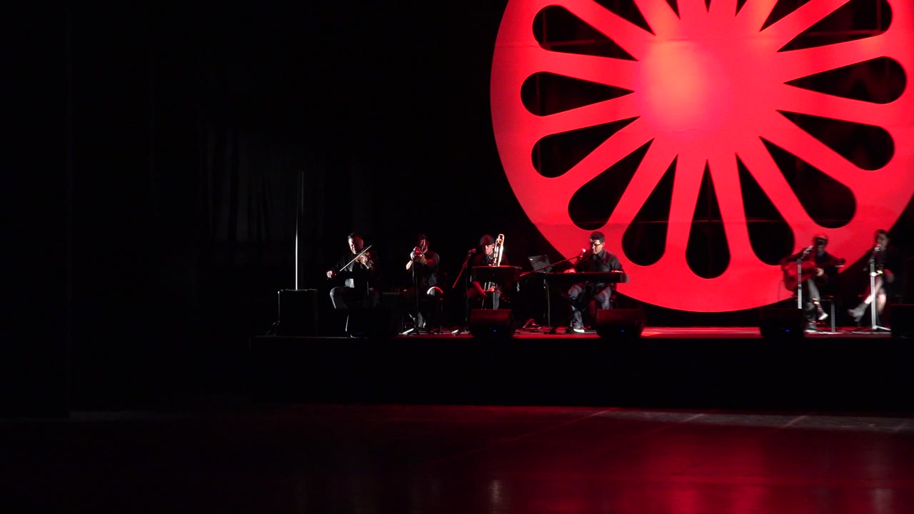 Wide stage frame from Cabo San Lucas, Mexico, with the ensemble spread across the lower frame and the red circular backdrop centered behind them.