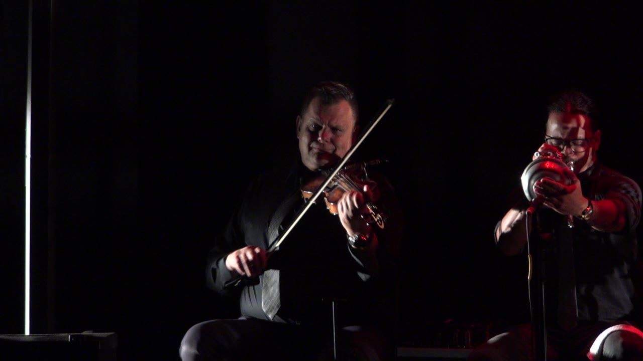 At the start of the Cabo San Lucas clip, an adult violinist sits at left while another adult musician faces forward with a trumpet under red stage light.