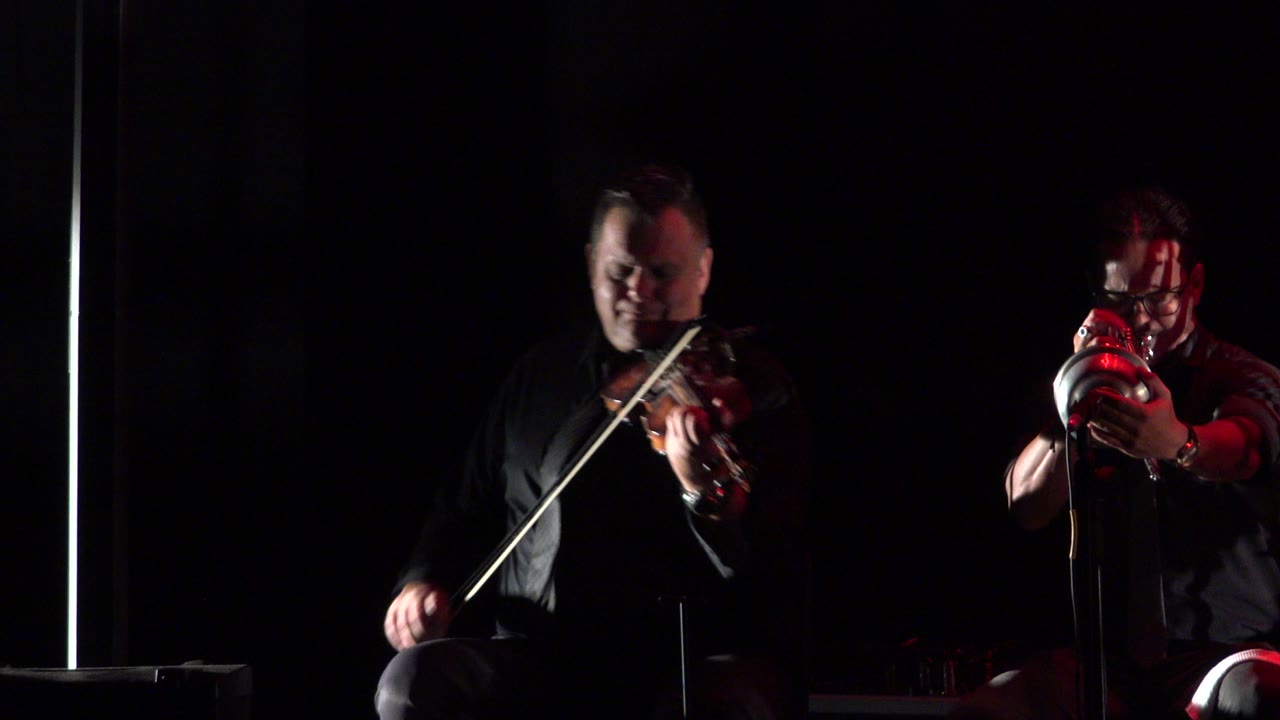 Seated violinist bows while a trumpet player faces the camera on stage.