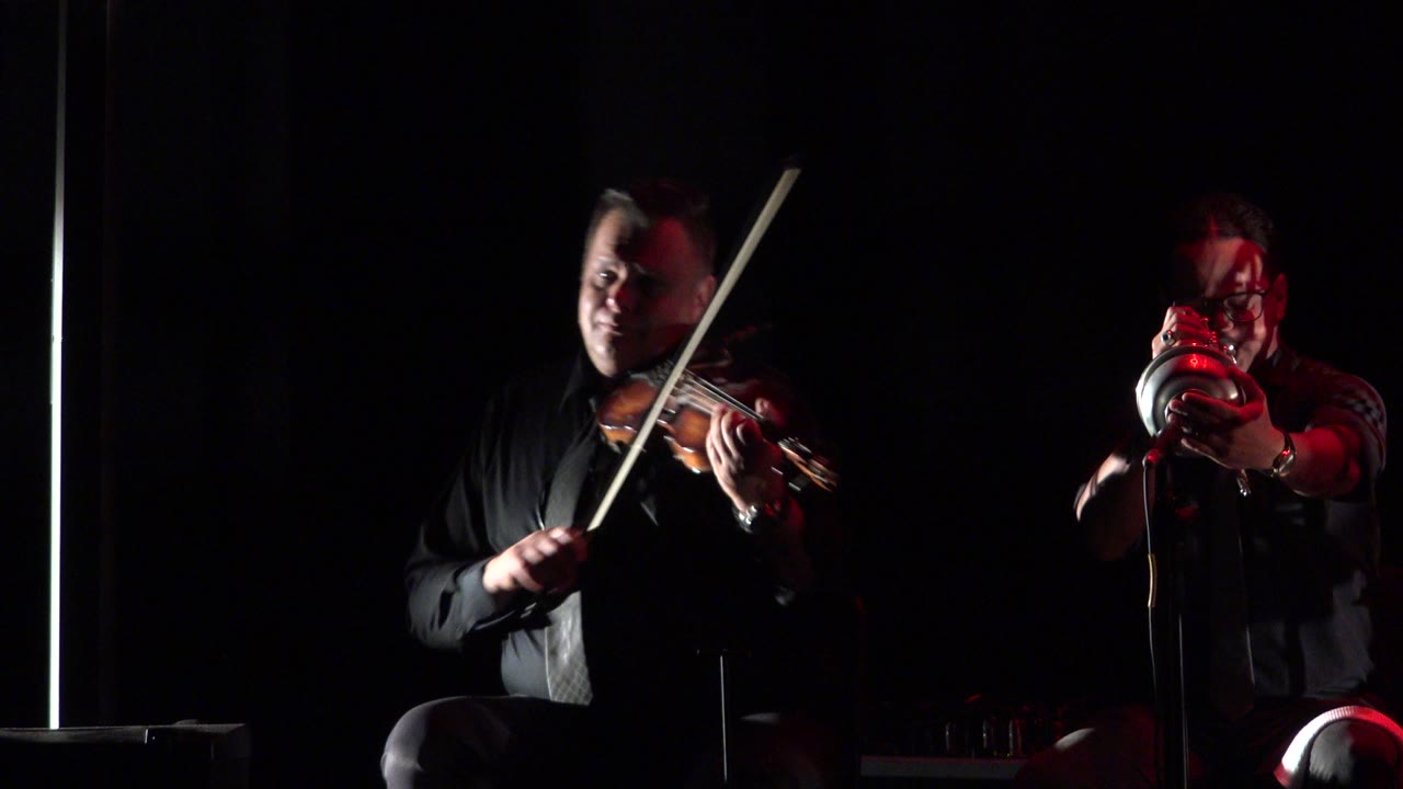 Two seated musicians perform on a dark stage.
