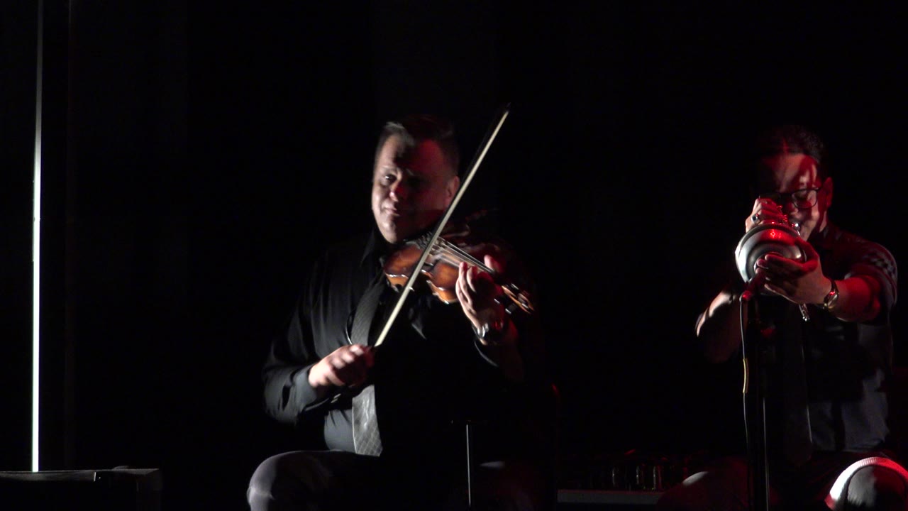 The violinist holds his bow across the strings while the brass player faces the camera under red light on the dark Cabo San Lucas stage.