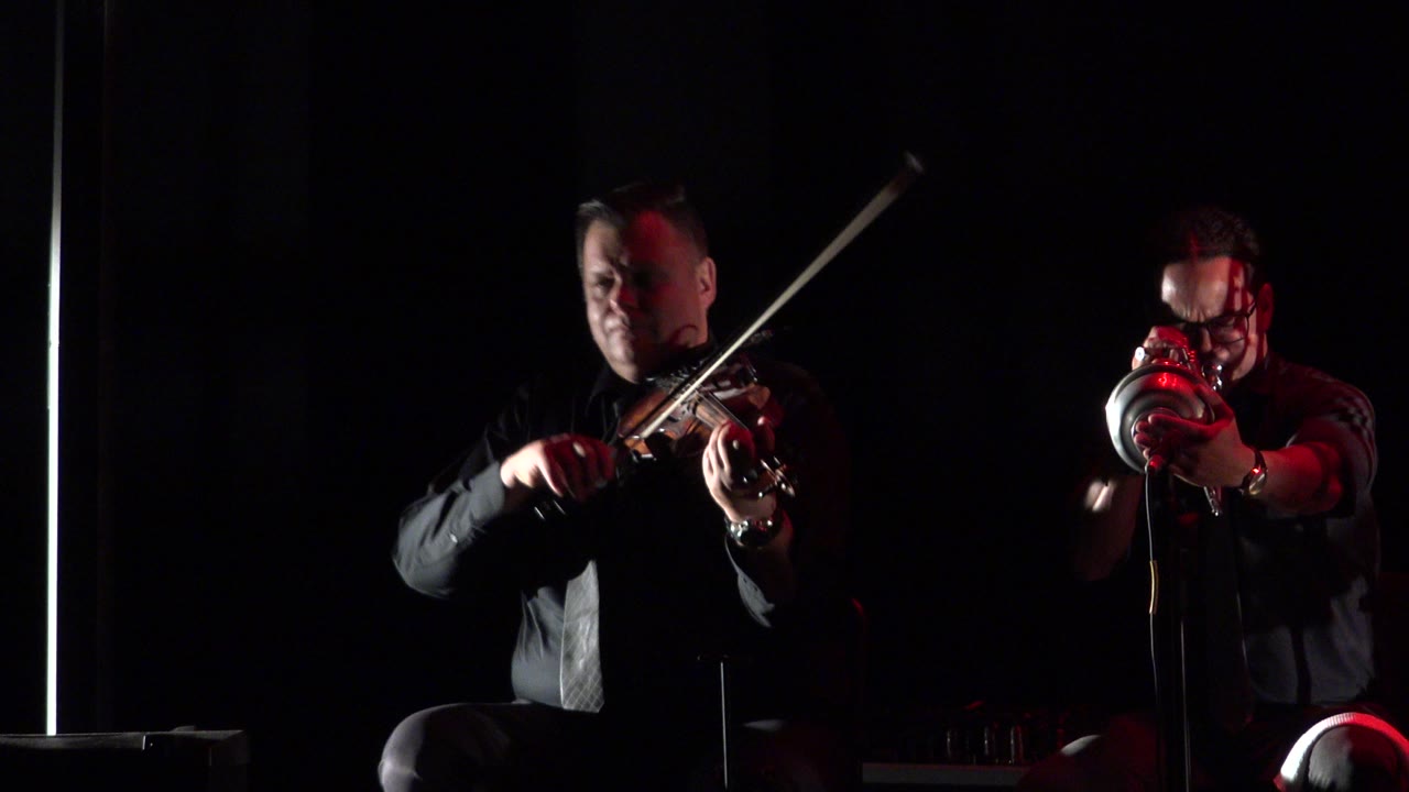 In Cabo San Lucas, Mexico, the violinist remains brightly lit on the left as the trumpet player on the right points the instrument forward under red lighting.