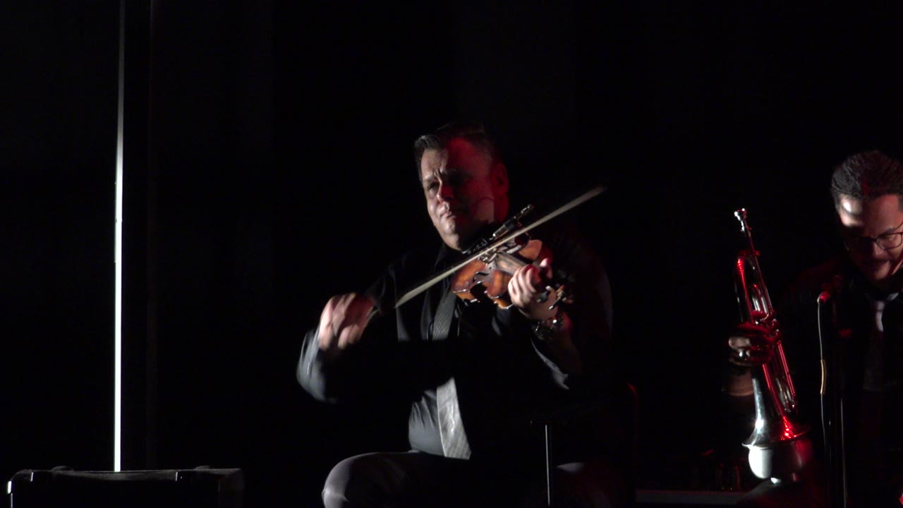 A violinist performs on a dark stage in Cabo San Lucas, Mexico, with a second musician partly visible holding a red brass instrument at the right edge.
