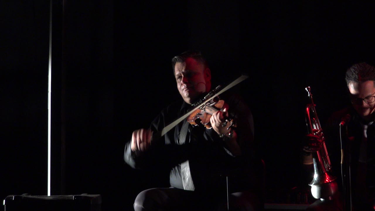 The Cabo San Lucas stage shot holds on the violinist as his bow arm moves, while the second musician remains in shadow beside a red brass instrument.