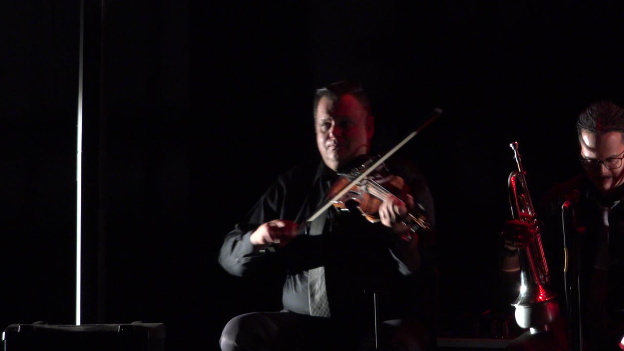 Violinist playing near center frame with red light on the adjacent musician.