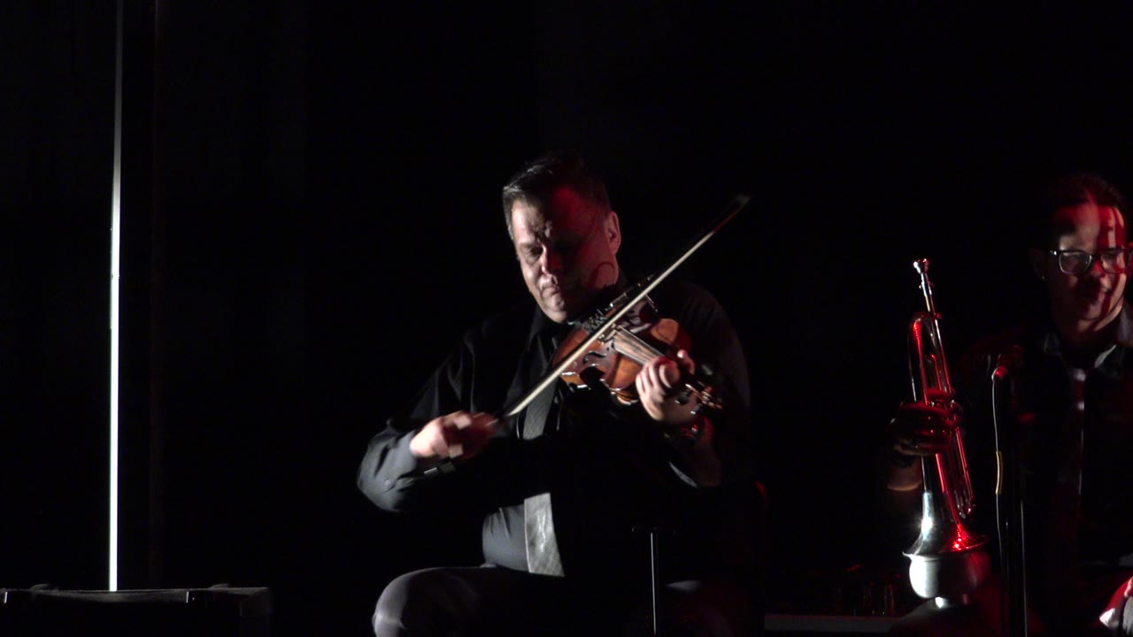 In Cabo San Lucas, Mexico, the violinist bows across the instrument while the trumpet player remains behind him, lit by a narrow red highlight.