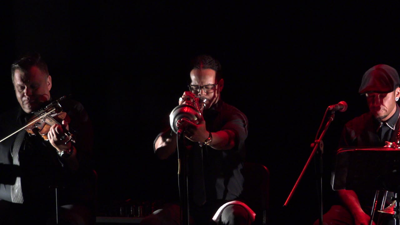 Three adult male musicians perform on a dark stage in Cabo San Lucas, Mexico, with a violinist at left, a brass player centered, and a seated trombone player near a microphone at right.