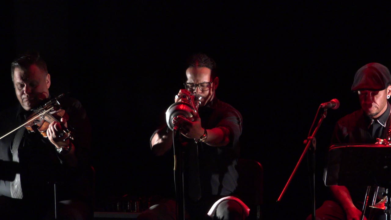 In Cabo San Lucas, Mexico, the violinist shifts farther into view at left while the center musician points a brass instrument toward the camera and the seated trombone player remains at right.