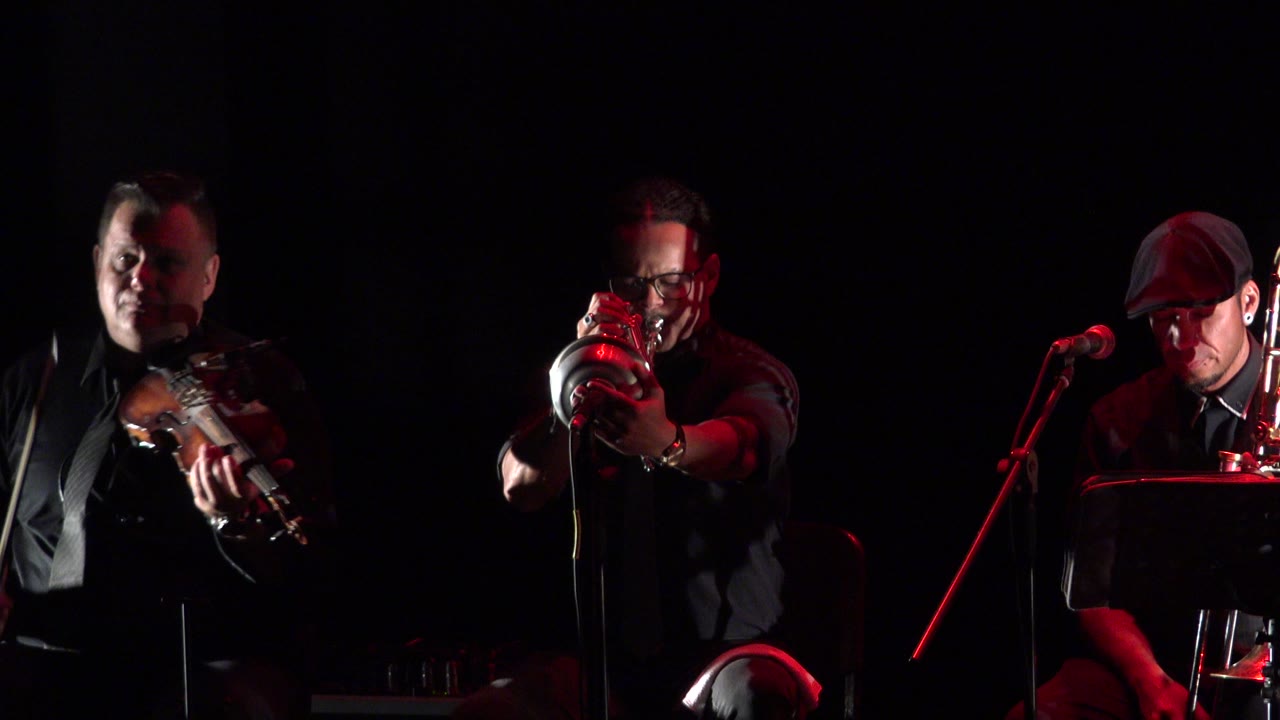 Center brass player and seated trombone player under red light.