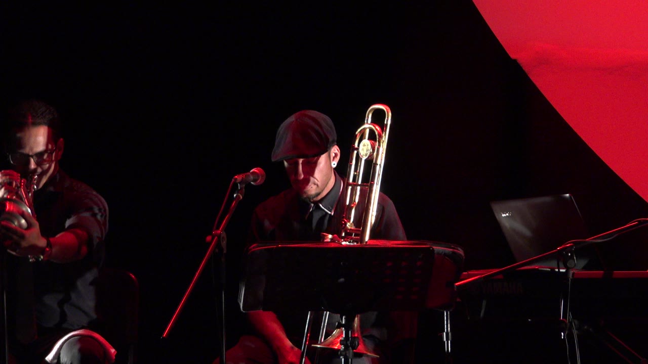 Trumpet player and trombone player on a dark red-lit stage.
