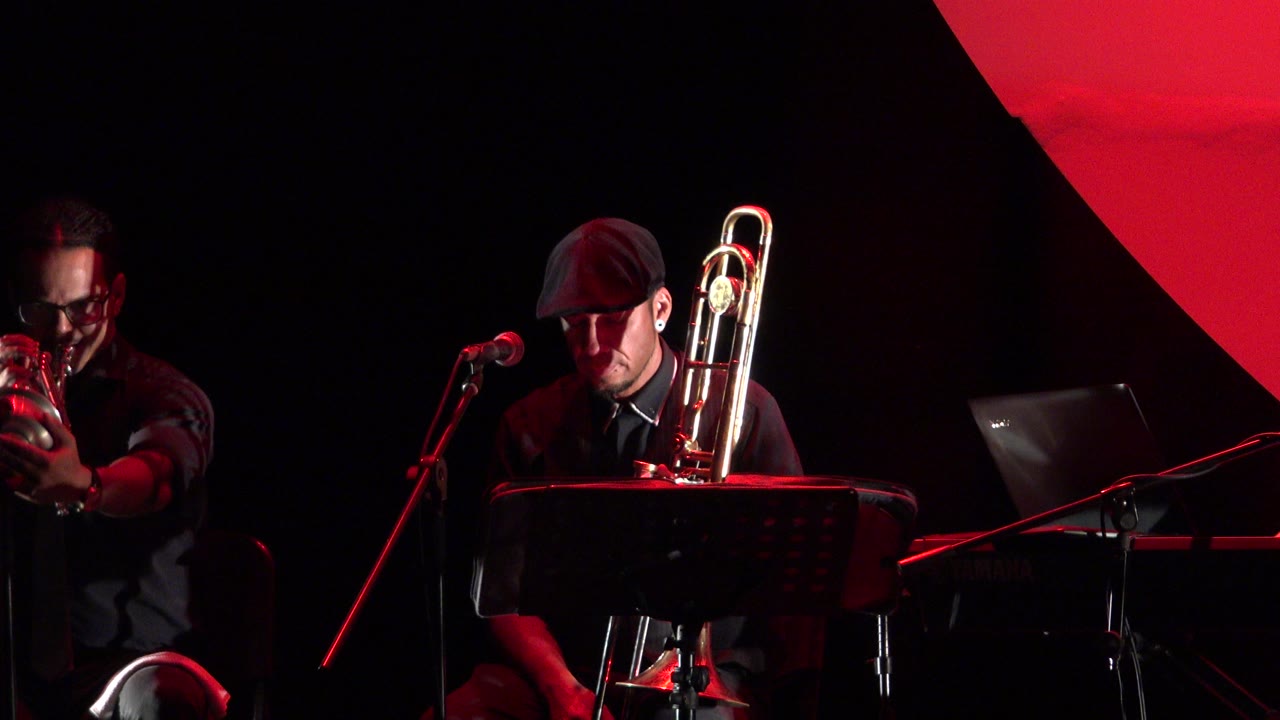 At 0:01 in Cabo San Lucas, the trumpet player remains at the left edge and the trombone player sits near center with his instrument upright beside a microphone.
