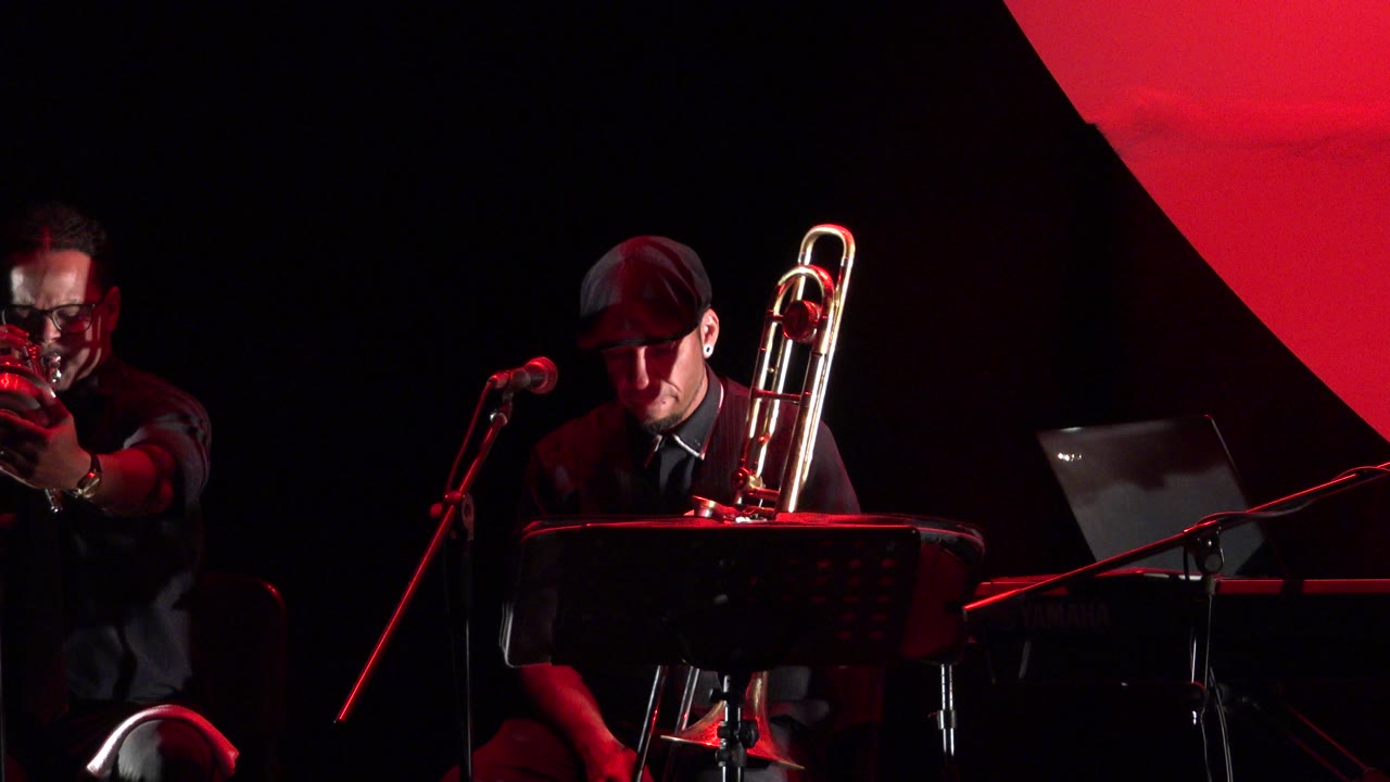 Jazz musicians on a dark red-lit stage with trombone, trumpet, keyboard, and microphones.