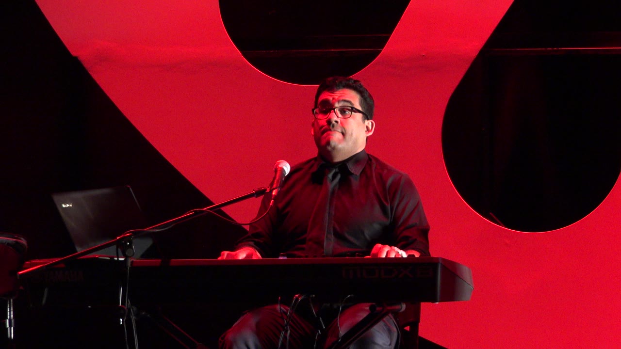 At 0:00, a man in glasses and a black shirt plays keyboard at a microphone on a red-lit stage in Cabo San Lucas, Mexico.