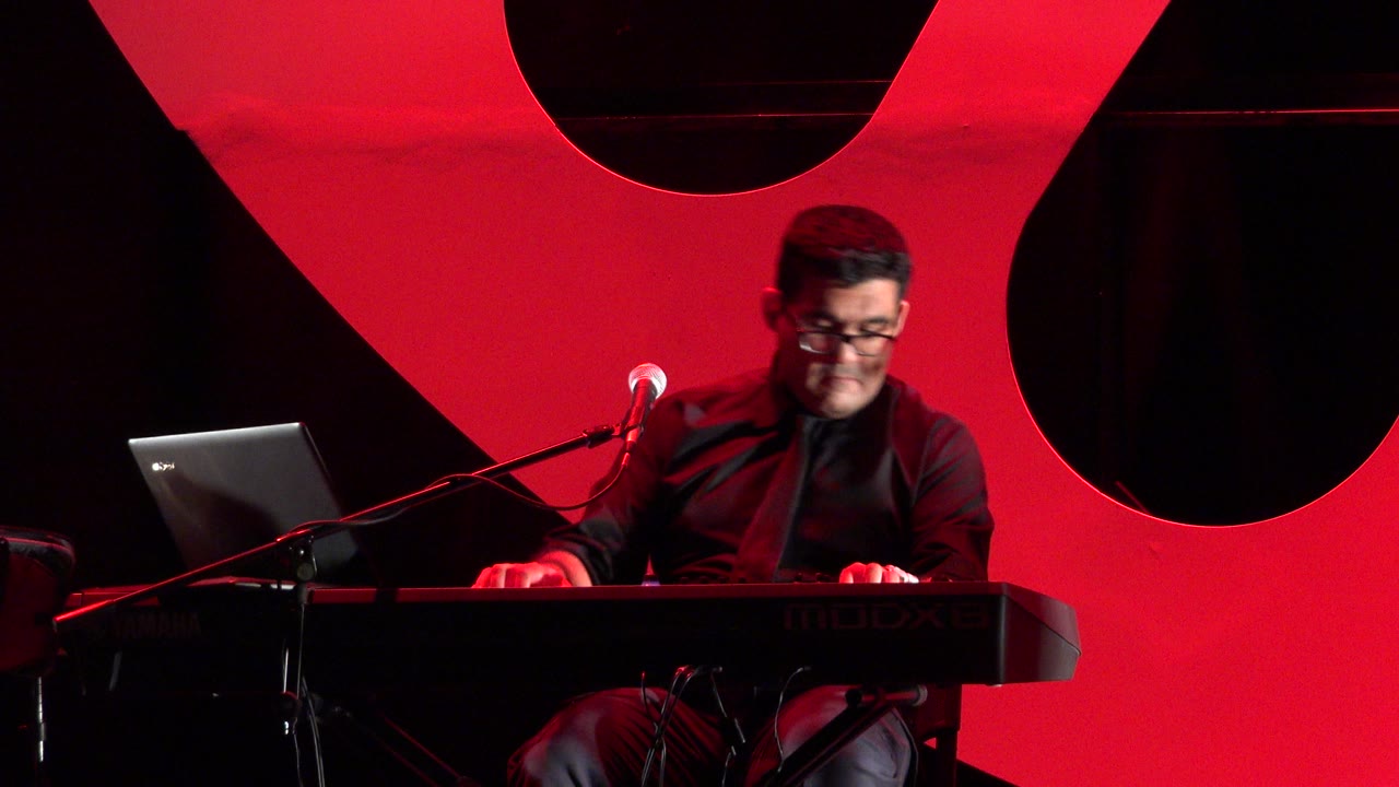 Man playing keyboard on a red-lit stage beside a microphone and laptop.