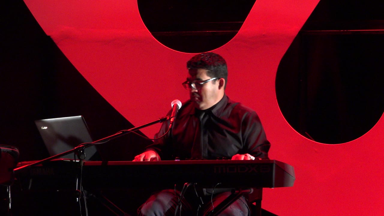 Musician at a keyboard with a laptop and microphone on a red-lit stage.