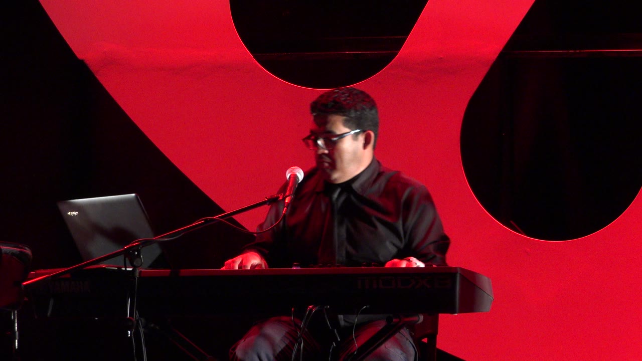 The Cabo San Lucas stage performance continues as the keyboard player faces the microphone, with his hands near the synthesizer and the laptop screen angled toward him.