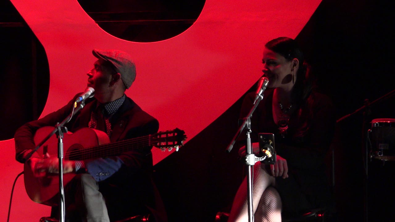 Two adult performers sit at microphones during a Cabo San Lucas pop rock show, with a guitar at left and a large red stage panel behind them.