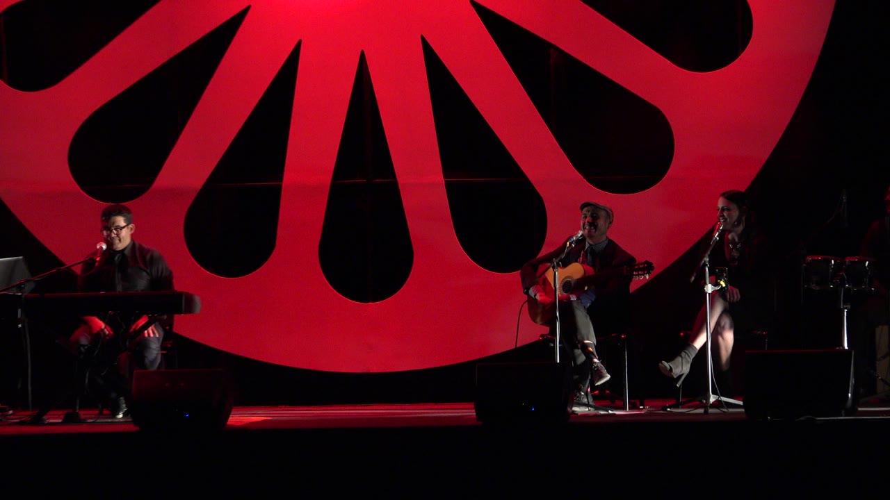 The stage remains in a wide view as the Cabo San Lucas band plays under red light, with the keyboardist and guitarist more clearly separated across the lower frame.