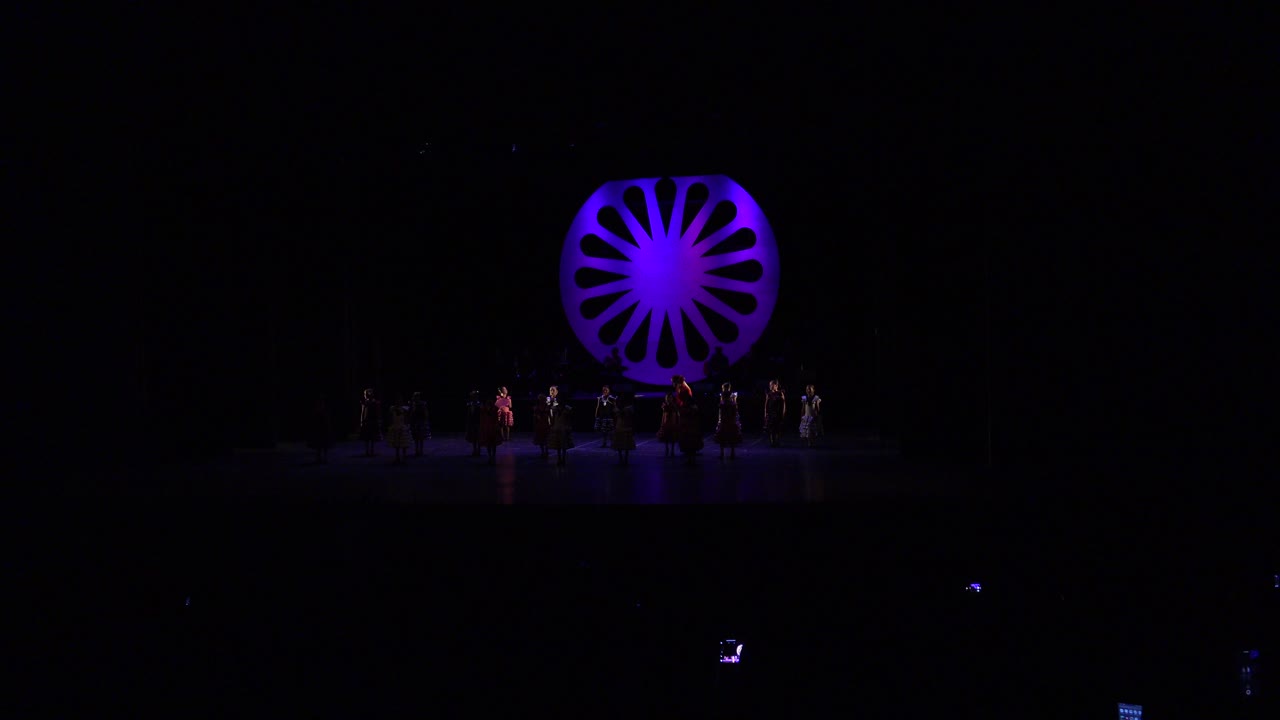 Wide audience-view frame of girls on a Cabo San Lucas stage beneath a purple circular illuminated backdrop, with musicians dimly visible behind them.