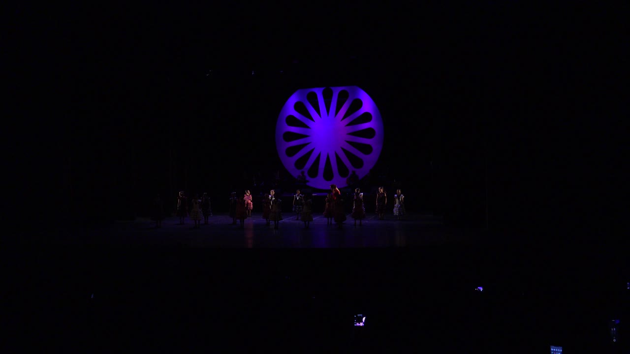 Wide frame of the Cabo San Lucas performance with girls spread across the stage and a bright purple circular set piece glowing behind the group.