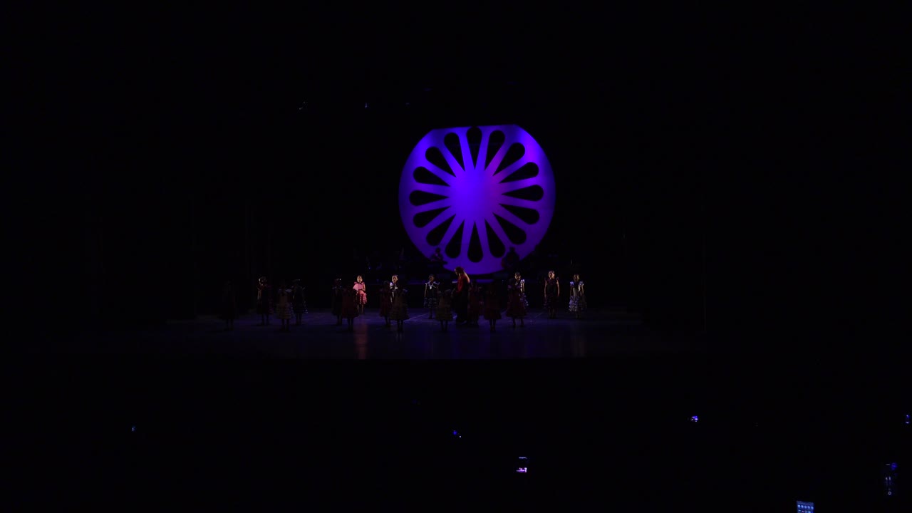 Wide stage scene with dancers and a purple circular backdrop.