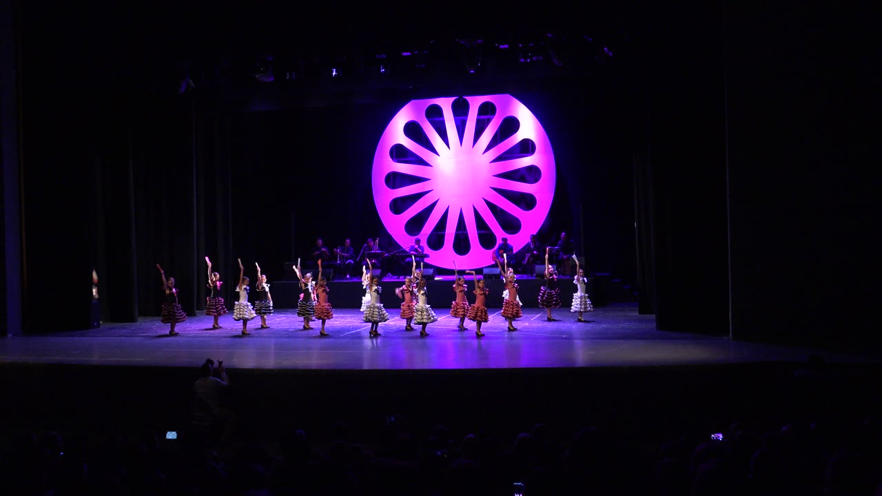 Wide view of young girls dancing on a Cabo San Lucas stage beneath purple light, with a bright magenta circular set piece centered behind them.