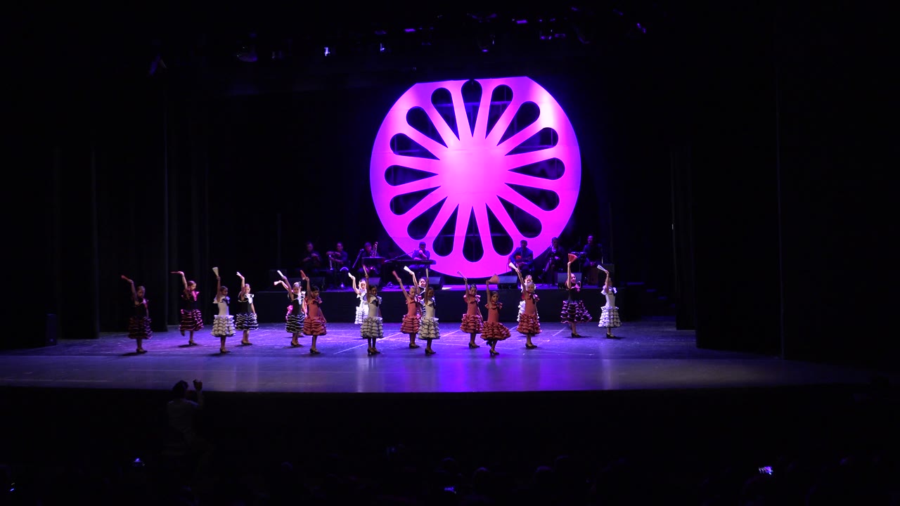 The dancers move farther across the Cabo San Lucas stage, with the magenta circular backdrop still centered and the audience area mostly black below.