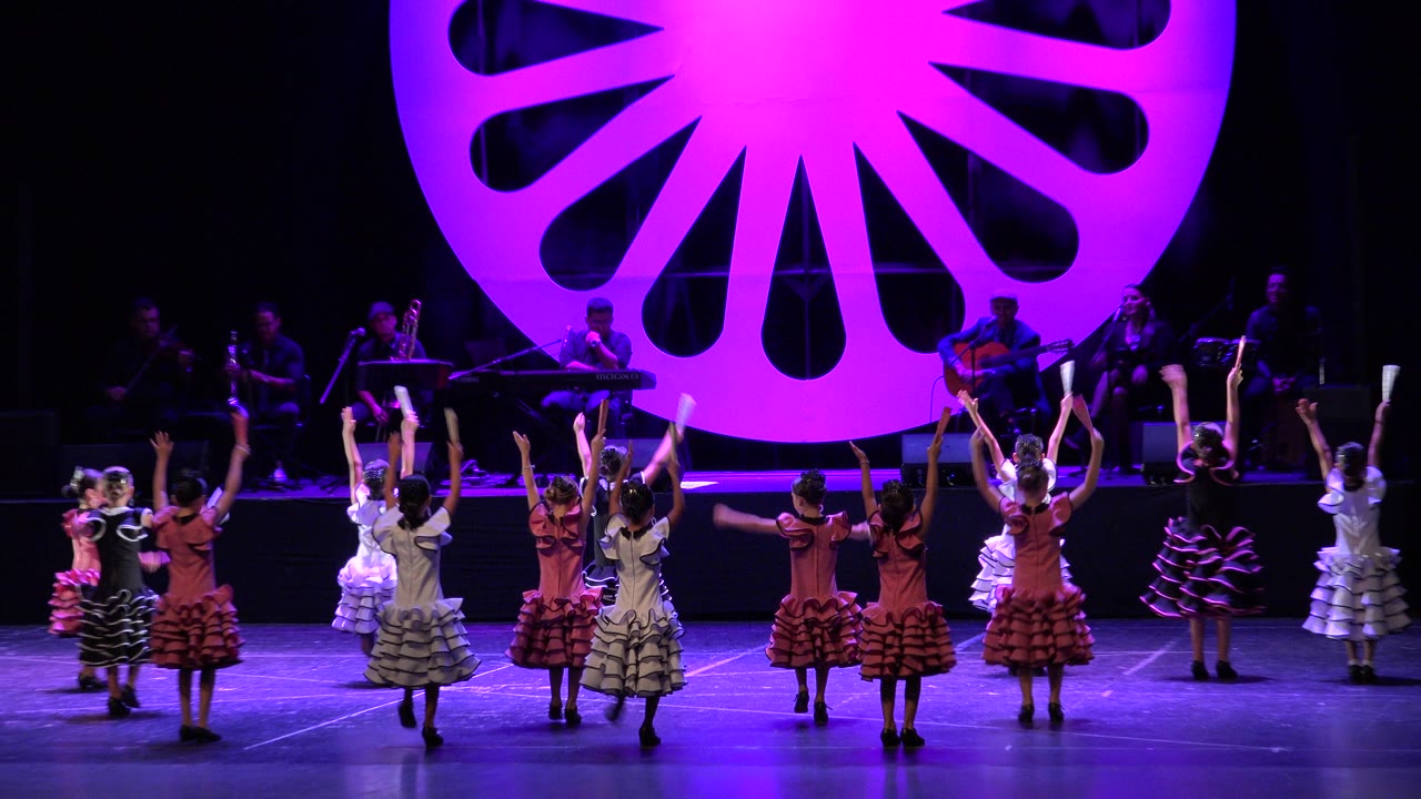 Children perform in rows on a purple-lit stage in Cabo San Lucas, Mexico, with musicians seated in shadow behind a large magenta fan-shaped backdrop.
