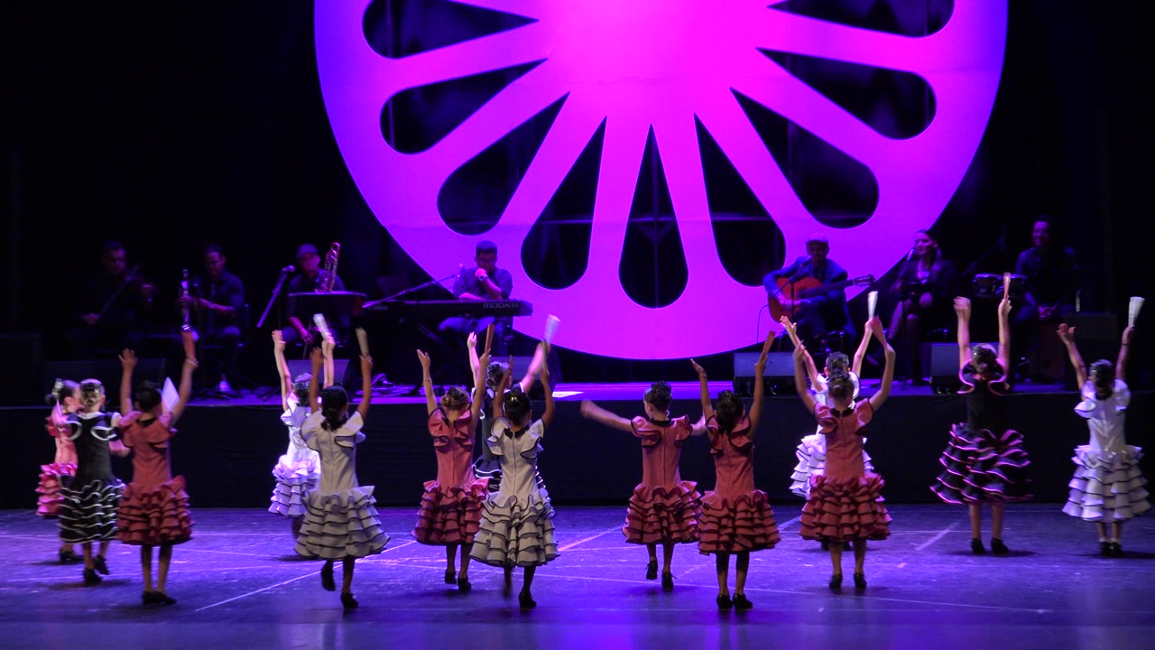 Children in pink, white, black, and patterned dresses raise their arms during a coordinated dance on a Cabo San Lucas stage with musicians in the background.