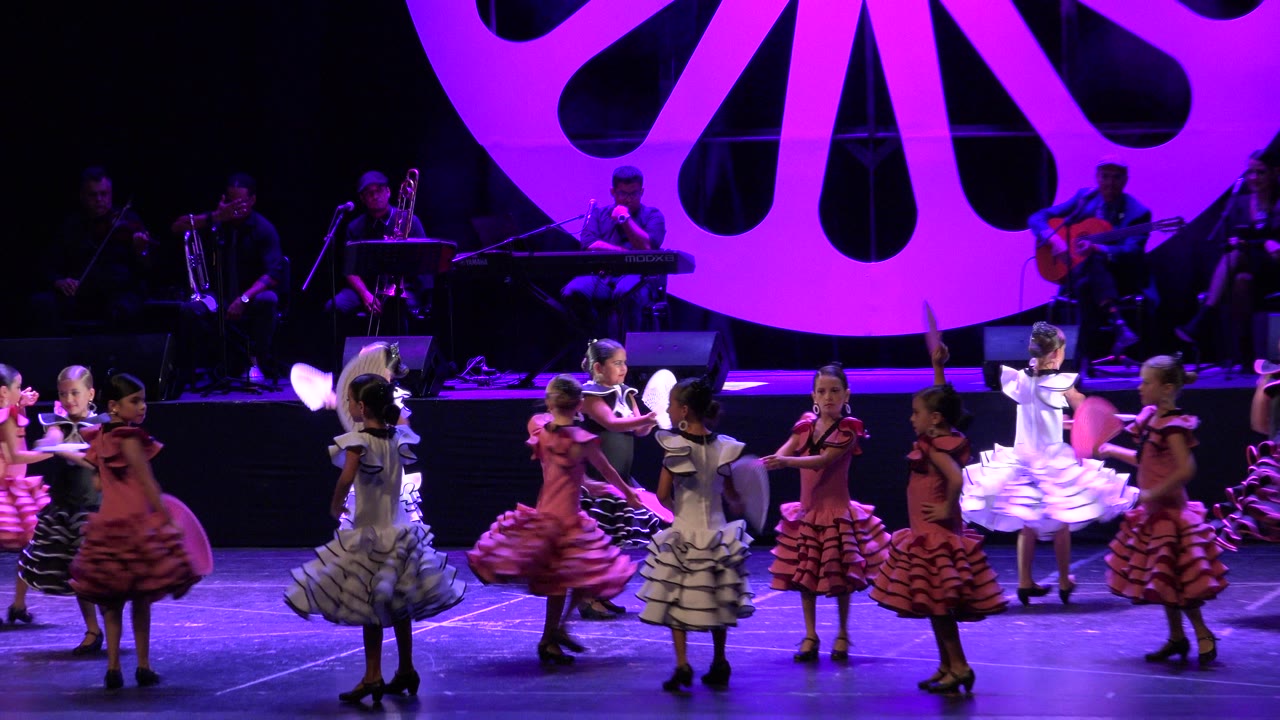 Children in ruffled dance costumes perform with fans on a purple-lit stage.