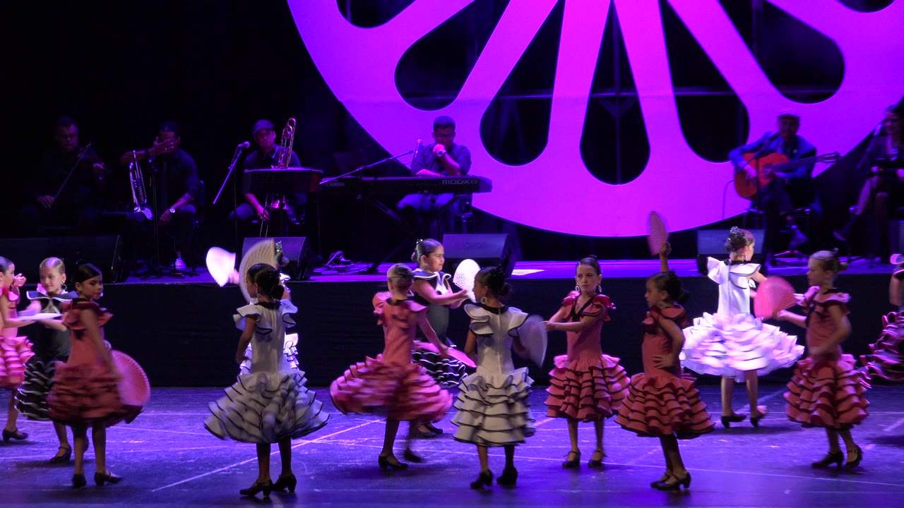 A line of children performs a fan dance in Cabo San Lucas, Mexico, with raised arms, ruffled skirts, and a seated band set against the large purple stage decoration.
