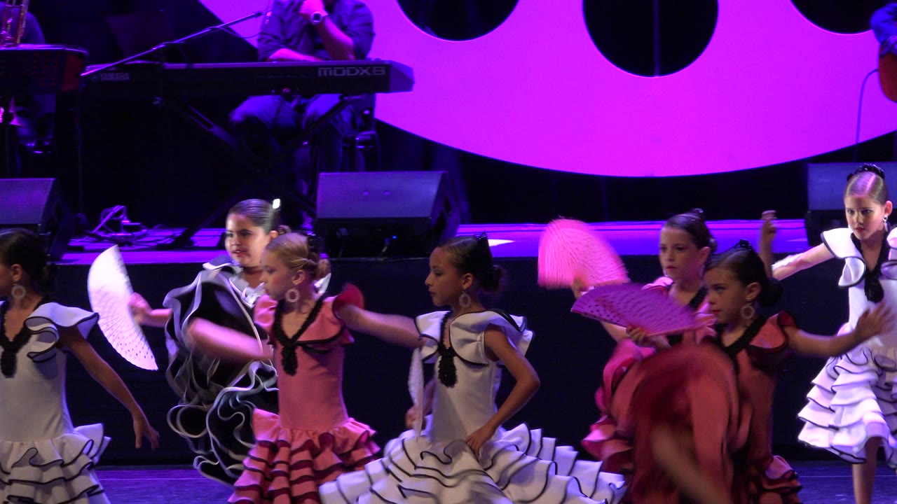 Young girls in ruffled dance costumes perform with fans on a pink-lit stage.