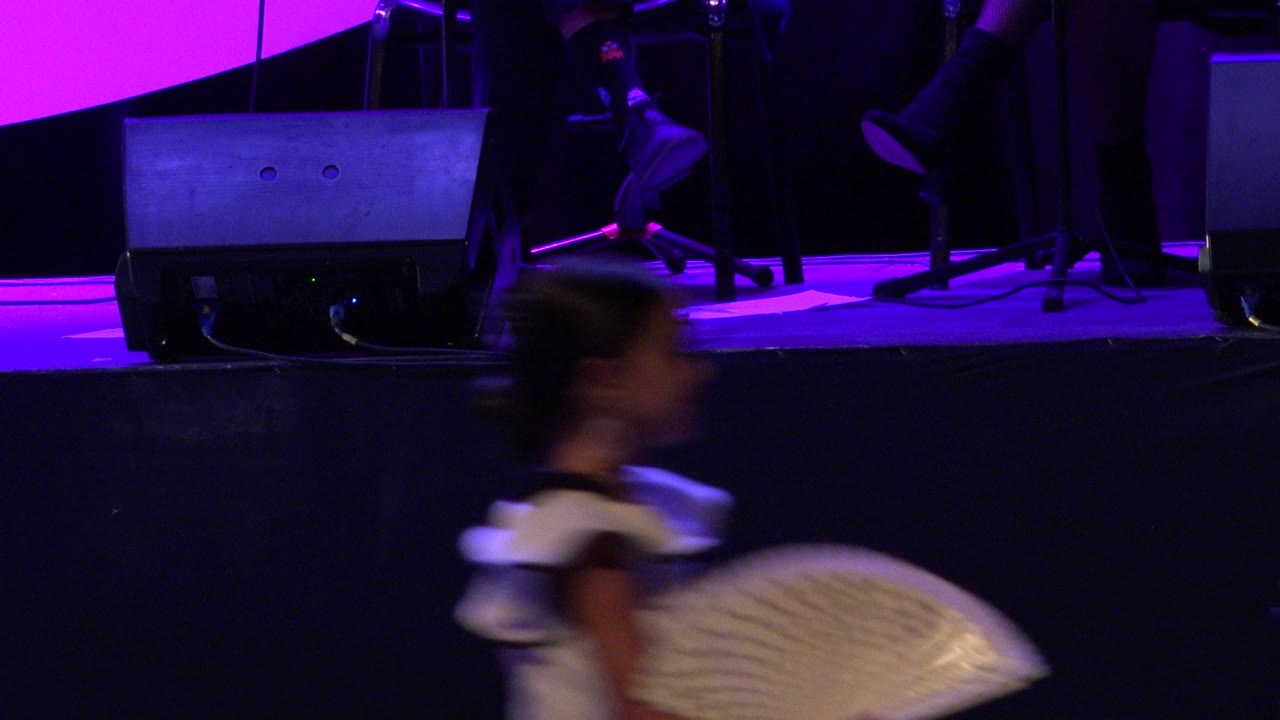 Blurred girl in a white dress carrying a fan below a purple-lit stage.
