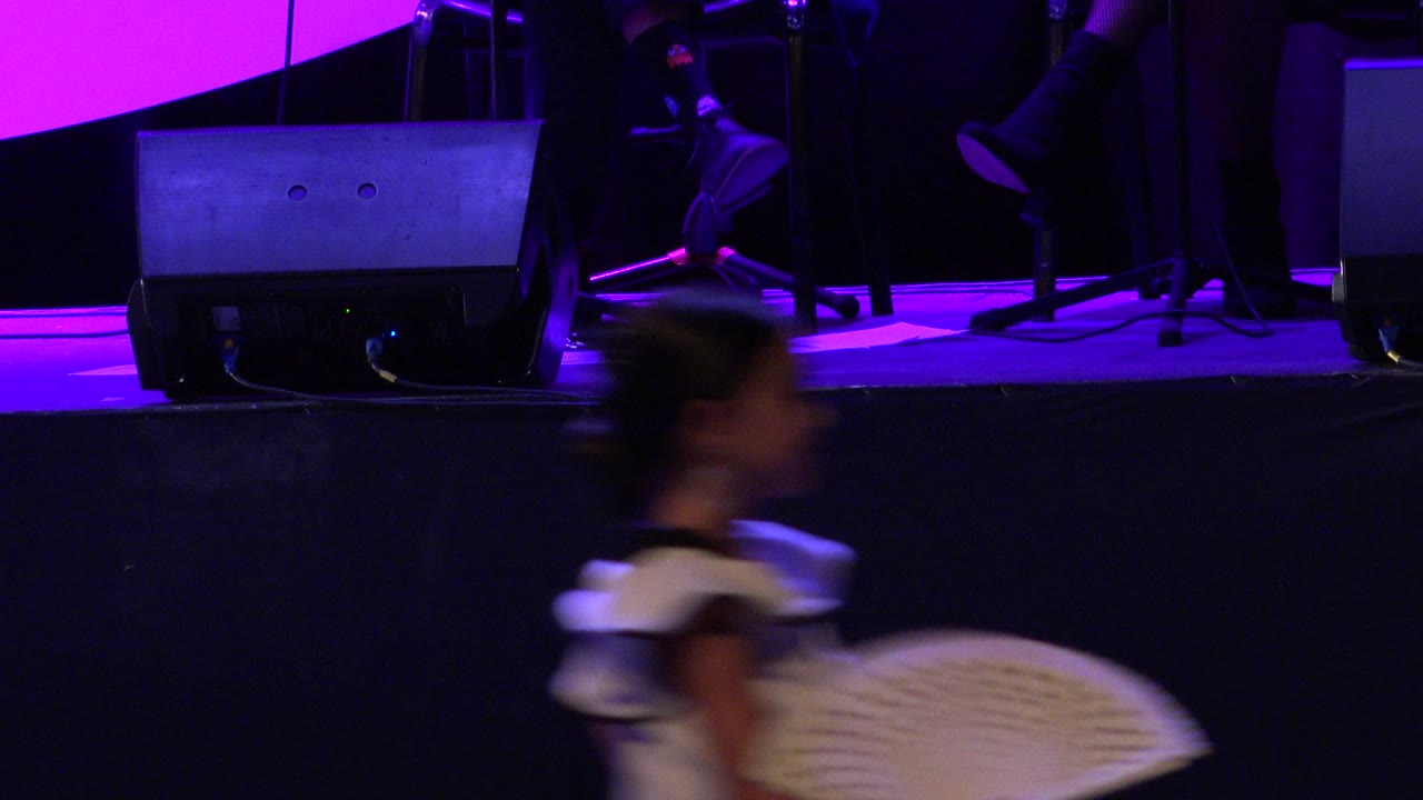 At 0:01 in Cabo San Lucas, Mexico, a child dancer crosses the lower frame with a pale fan, partially blurred against purple lighting and black stage equipment.