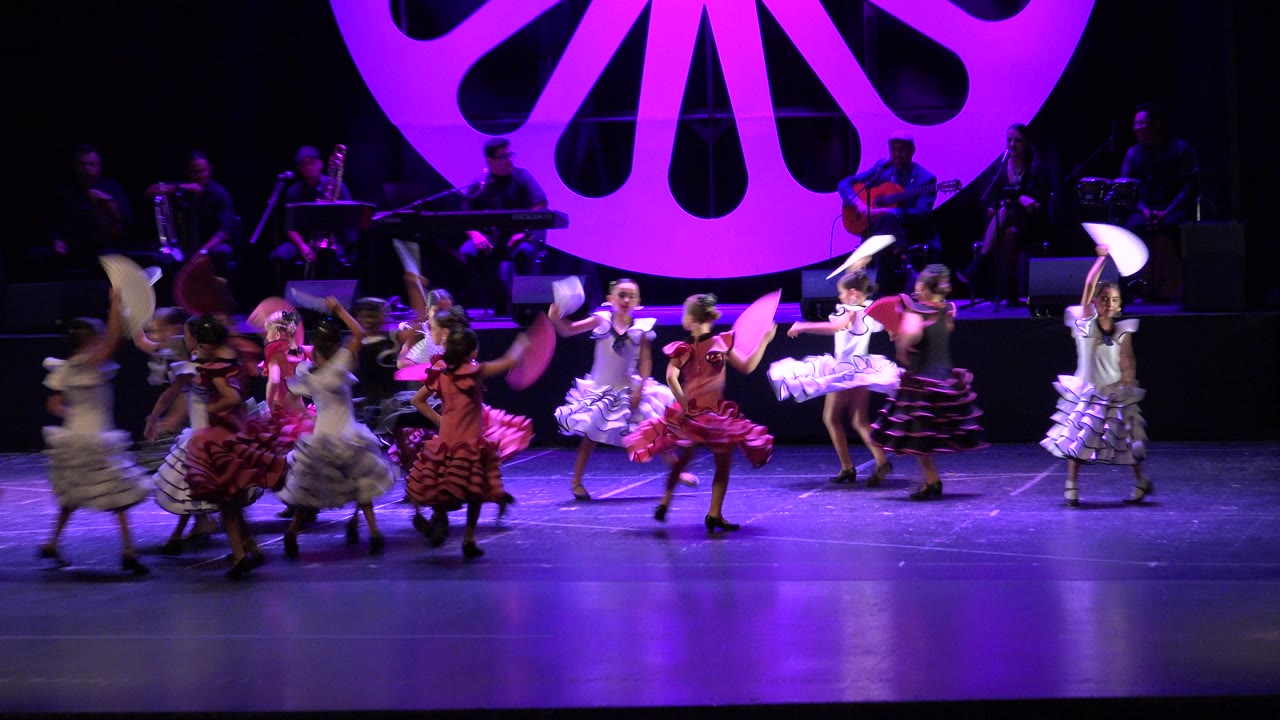 Children perform a choreographed dance on a purple-lit stage in Cabo San Lucas, Mexico, with musicians and a large shell-shaped backdrop behind them.