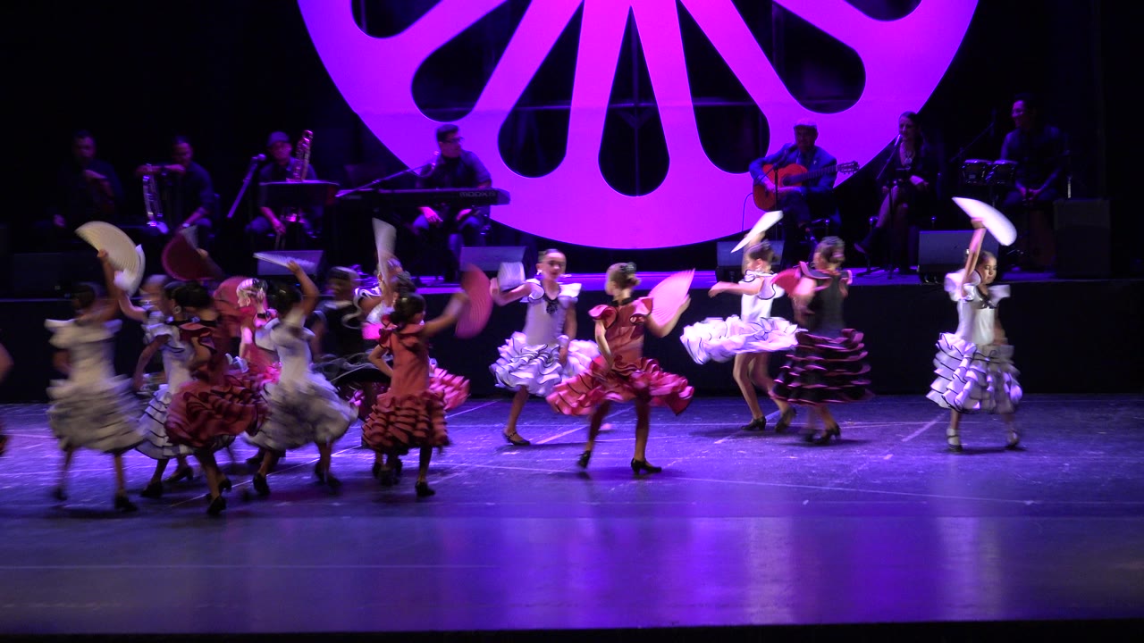 Girls in pink, white, red, and dark ruffled costumes dance in formation with fans while a band sits in shadow at the rear of the Cabo San Lucas stage.