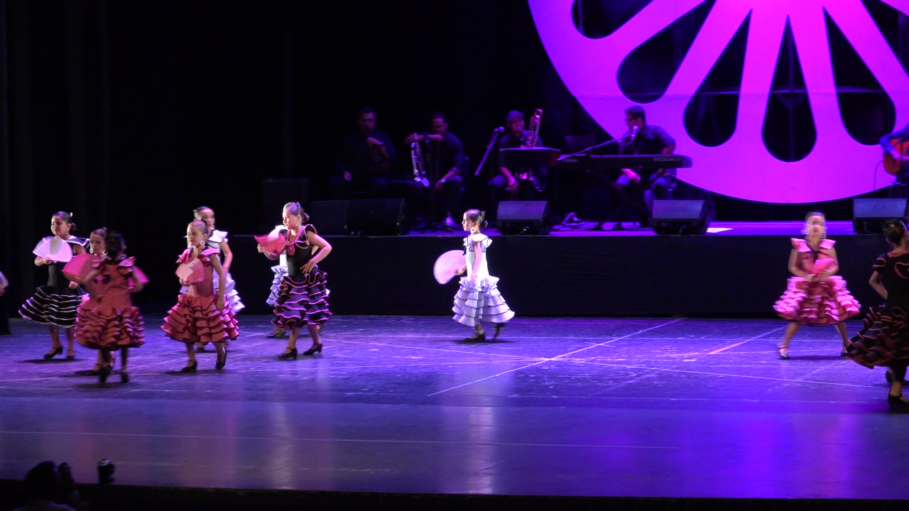 Young girls in ruffled dresses dancing on a purple-lit stage.
