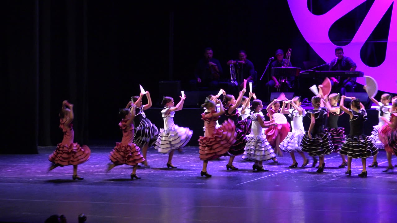 Wide view in Cabo San Lucas, Mexico, of young girls in ruffled dresses dancing across a purple-lit stage while musicians sit in the background.