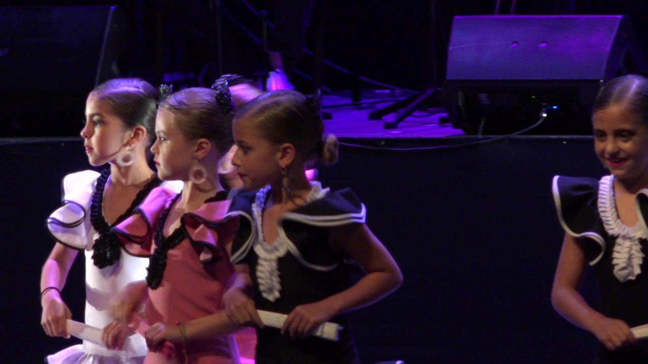 Costumed girls stand on a purple-lit stage holding white rods.