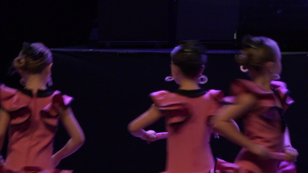 In Cabo San Lucas, three children in pink ruffled dance outfits shift positions on a dark stage while purple light catches their shoulders and hair.