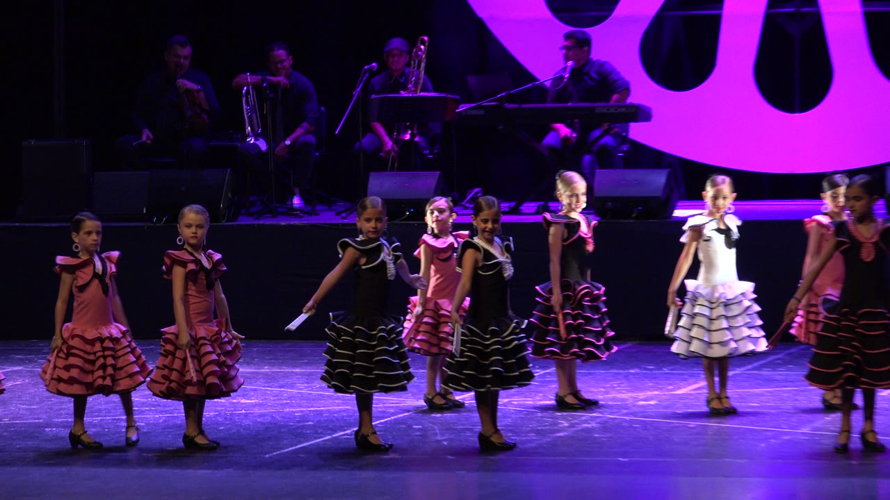 Young girls in ruffled dresses dance on a purple-lit stage.