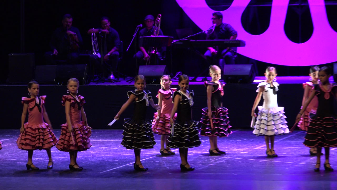 Children in ruffled performance dresses continue their line dance on the Cabo San Lucas stage while the musicians remain seated behind them.