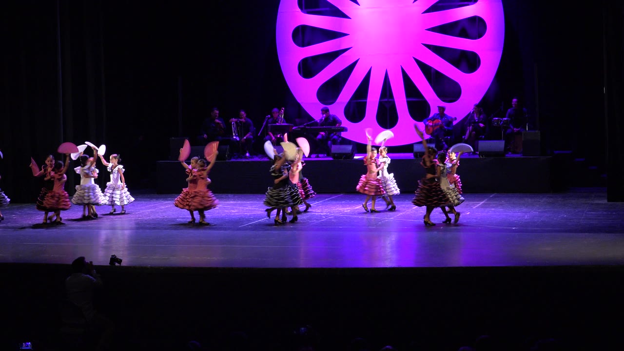 Children in ruffled dresses dance in small groups on the Cabo San Lucas stage while white and pink fans are lifted at different angles beneath purple lighting.