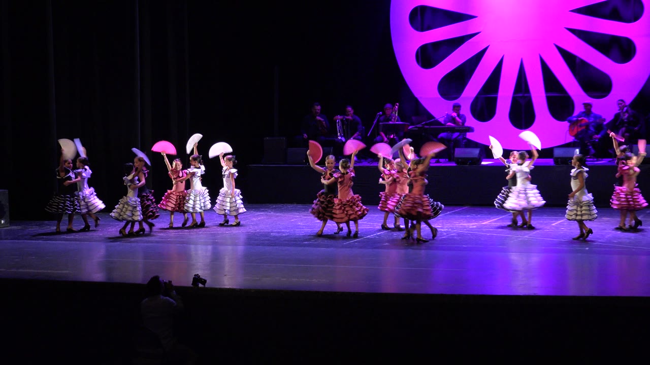 Children in pink, white, black, and striped costumes hold open fans while moving in small groups on the Cabo San Lucas recital stage.