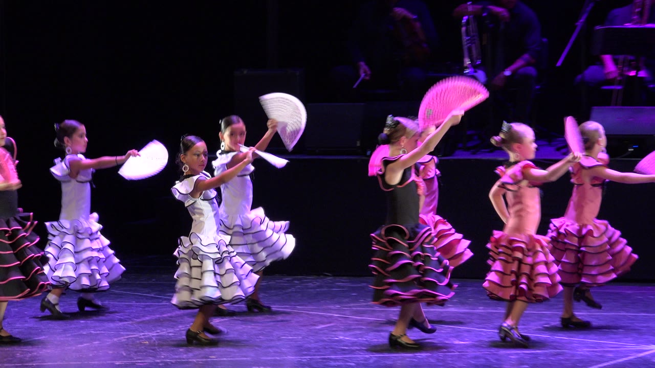 Young dancers in frilly dresses hold fans on a purple-lit stage.
