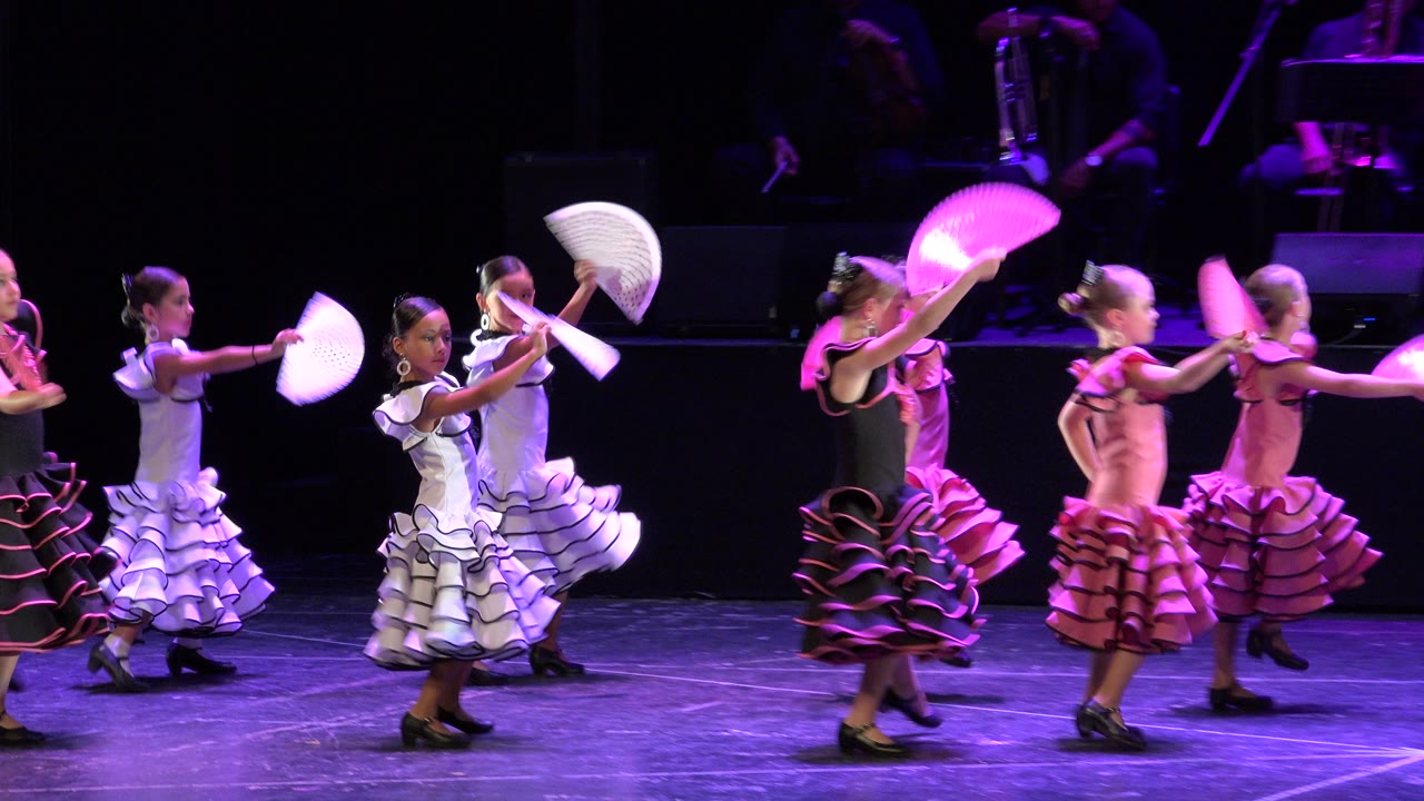 Children in white, pink, and black dresses extend their fans in a line as they cross the theater stage in Cabo San Lucas, Mexico.