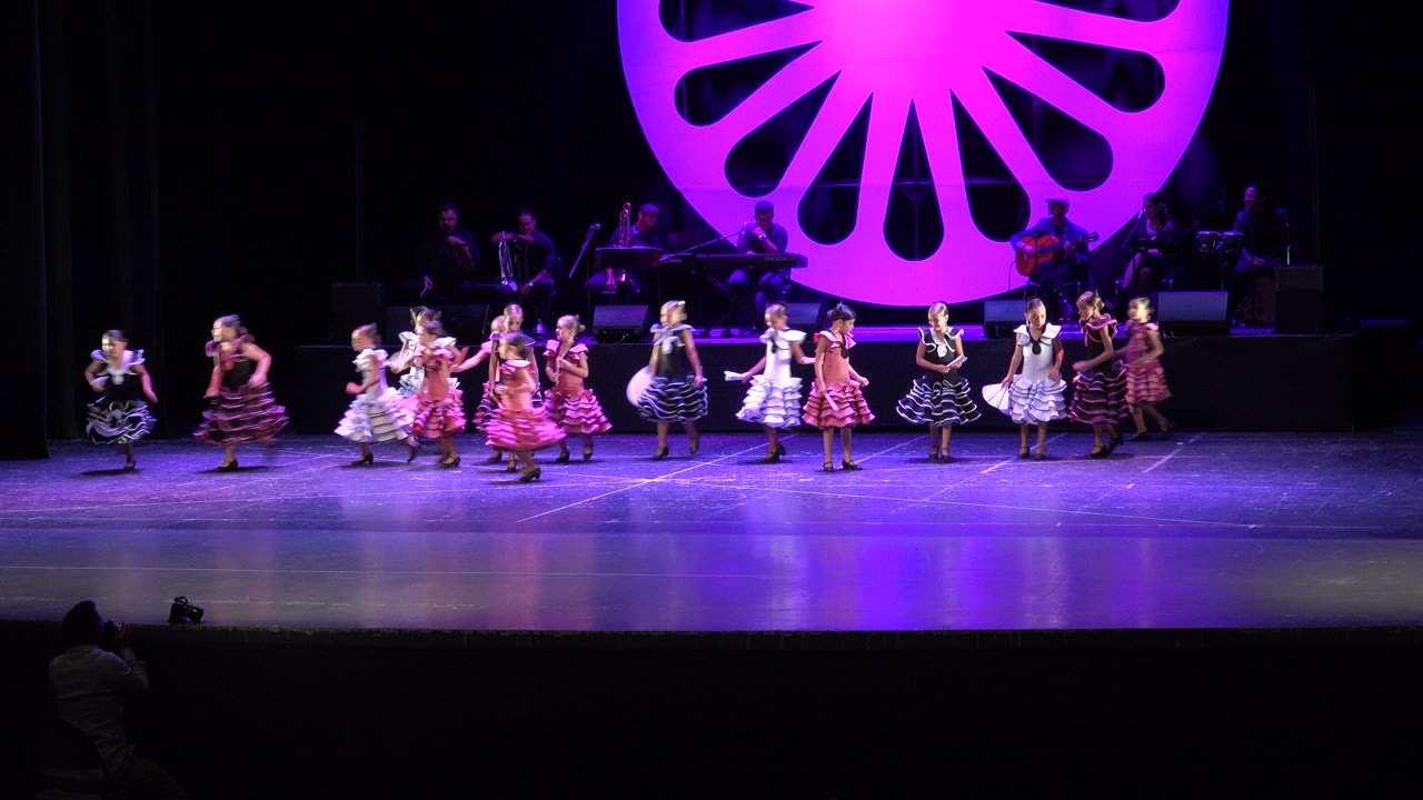 At 0:00 in Cabo San Lucas, girls in colorful ruffled dresses spread across a theater stage under purple lighting with a large magenta fan-shaped set piece behind them.