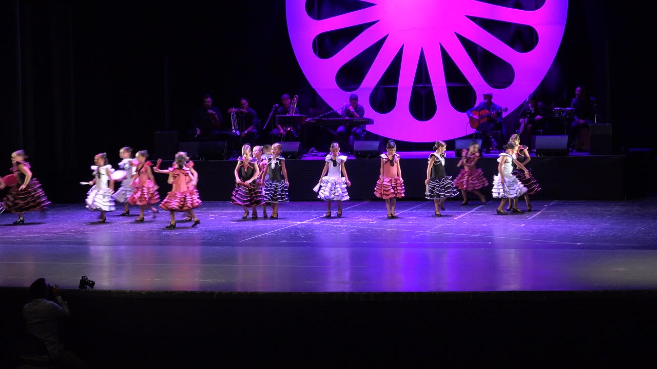 Girls in dresses form a looser line on stage under purple lighting.