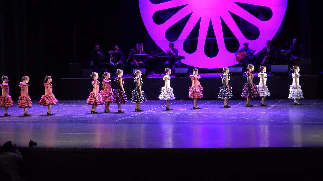 At 0:01, the girls hold their places in a wide stage formation while the purple lighting reflects on the floor and the background musicians remain partly visible.