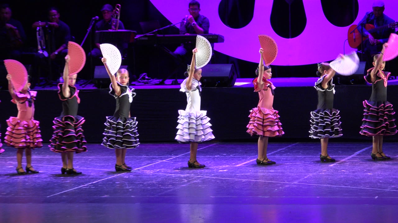 Children in pink, black, and white ruffled dresses stand in a line on a purple-lit stage in Cabo San Lucas, Mexico, with musicians visible behind them.